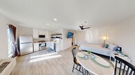 This well-designed single-floor unit makes excellent use of its compact footprint through a thoughtful open-concept layout that seamlessly integrates the kitchen, dining, and sleeping areas into one bright, cohesive space. Light wood-look tile flooring runs throughout, lending a clean and airy feel that is complemented by crisp white walls and recessed lighting.

The kitchen is a functional highlight, featuring white shaker-style cabinetry with gold hardware, a marble-style tile backsplash, light quartz countertops, and a full suite of stainless steel appliances including a gas range, over-the-range microwave, refrigerator, and dishwasher. Open dark wood shelving adds a modern contrast and additional storage.

The open living and sleeping area is bright with natural light from a front-facing window and benefits from a ceiling fan for added comfort. A wall-mounted flat-screen television and a compact dining set with a round table and two chairs define the living zone, while the bedroom area accommodates a full-size bed and nightstand.

The bathroom features a pedestal sink beneath a gold-framed vanity mirror, a shower with curtain, and a stylish three-bulb light fixture. A dedicated laundry area and a wall-mounted HVAC unit round out the practical amenities of this efficiently appointed unit.