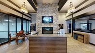 Located at 100 Hawk Ridge Dr in Iowa City, Iowa, this single-floor leasing and amenity space for Hawks Ridge offers a well-appointed, modern environment designed to welcome residents and prospective tenants alike. The main lobby serves as the heart of the space, anchored by a dramatic floor-to-ceiling stone fireplace topped with a wall-mounted display. Exposed dark wood beam accents traverse the ceiling, complementing the warm stone tile flooring and pendant lighting to create an inviting, contemporary-rustic atmosphere. A front reception counter provides a functional and stylish welcome area, while large glass sliding doors offer a direct view into an adjacent sports court facility, flooding the space with natural light and visual openness. The dining and living area features a round wooden dining table with chairs, seamlessly connected to the reception zone. The kitchen is fully equipped with stainless steel appliances, dark espresso cabinetry, granite countertops, and a dedicated coffee bar station, reflecting thoughtful attention to resident convenience. A private office with hardwood flooring, a ceiling fan, a workstation, and interior windows offers a professional setting for leasing consultations. The entry vestibule features the Hawks Ridge branded entryway, reinforcing the community's identity from the moment guests arrive.