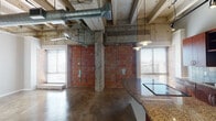This one-bedroom, one-bathroom unit in Houston's 77021 corridor showcases a distinct industrial aesthetic rooted in its original architectural character. Soaring ceilings with exposed concrete beams, visible ductwork, and structural columns create a raw, loft-like atmosphere throughout the open living area. Polished concrete floors run continuously across the space, complementing the exposed red brick walls that serve as a defining design element.

The kitchen is a functional centerpiece, featuring rich wood cabinetry, granite countertops, a tiled backsplash, and a curved island that provides additional prep and seating space. Pendant lighting suspended from the high ceiling illuminates the kitchen, while stacked in-unit laundry adds everyday convenience. Multiple windows fitted with blinds allow natural light to pour in, brightening the polished floors and contrasting warmly against the raw brick and concrete surfaces.

The bedroom offers a quieter retreat, with a recessed window ledge and continued polished concrete flooring. The bathroom features a full tub and shower combination with classic white tile and a bold black accent band. The overall ambiance balances urban industrial character with functional, modern finishes throughout.