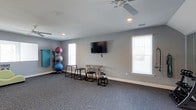 This single-floor fitness space offers a clean, functional environment designed for versatile exercise and movement. The room is finished with durable, textured carpet in a neutral gray tone, providing cushioning underfoot during workouts. Light gray walls contribute to an open, airy atmosphere, while white baseboards add a crisp, polished contrast throughout.

Natural light filters in through multiple windows fitted with horizontal blinds, offering brightness and ventilation control. A ceiling fan with a light fixture is centrally mounted, ensuring comfortable airflow during physical activity. Recessed lighting supplements the natural light, keeping the space well-illuminated at all times.

A wall-mounted flat-screen television serves as a focal point along one wall, ideal for following workout videos or entertainment during training sessions. The room is equipped with a stacked set of exercise balls in varying sizes, a resistance band rack, and a small collection of dumbbells. Several minimalist black cube step stools and a wooden bench provide options for step training, stretching, and seated exercises.

Overall, the space presents a well-organized, distraction-free fitness environment suited for individual training and light group exercise.