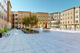 Located at 305 E Front St in downtown Missoula, Montana, this expansive open-air courtyard serves as a central gathering space within a modern multi-story building complex. The courtyard is defined by a uniform large-format paver surface that spans a generous footprint, creating a versatile open area suited for both events and everyday use. The surrounding building facades feature a contemporary mix of warm cedar-toned cladding, stone masonry, and stucco finishes, rising several stories on all sides to create an enclosed yet open-to-sky environment.

A standout architectural feature is a curved, glass-wrapped entrance pavilion anchoring one side of the courtyard, offering a striking focal point with floor-to-ceiling windows. Throughout the space, raised planter beds are filled with trees, shrubs, and ornamental grasses, adding greenery and texture to the hardscape. Metal planter boxes line the building perimeters, further softening the architectural edges.

Seating is distributed across the courtyard with benches, high-top tables with umbrellas, and a lounge area with chairs near the pergola — a rustic timber structure draped with climbing plants and strung with Edison-style bistro lights. A ping-pong table adds a recreational element. The open sky above ensures abundant natural light throughout the day, making this courtyard a vibrant, welcoming outdoor amenity.