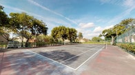 This outdoor recreational space features a full-size tennis court finished in a hard-surface playing surface with clearly marked white boundary lines and a center net. The court is fully enclosed by chain-link fencing, providing both safety and containment during play. Tall flood lights mounted on metal poles are positioned along the perimeter, enabling use during evening hours. The surrounding landscape is well-established, with mature trees forming a natural canopy backdrop and dense hedging lining portions of the fencing, lending a sense of privacy and greenery to the space. The open-air setting benefits from abundant natural light during daytime, with views of a partly cloudy sky adding to the pleasant outdoor atmosphere. Neighboring residential structures are visible beyond the tree line, indicating the court is situated within or adjacent to a residential community. The overall setting is functional and well-equipped for recreational tennis, combining practical infrastructure with a naturally landscaped environment that enhances the playing experience.
