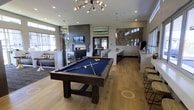 This single-floor amenity space is designed with a contemporary aesthetic and a welcoming, social atmosphere. The open-concept layout accommodates multiple activity zones seamlessly. At the forefront sits a full-sized billiards table with a blue felt surface, positioned beneath a striking sputnik-style chandelier that serves as a central design focal point. Recessed lighting throughout complements the natural light filtering in from windows along the perimeter.

The lounge area features a modern fireplace framed in sleek dark tile, flanked by a wall-mounted television, creating a comfortable gathering spot. A seating arrangement is positioned nearby, encouraging relaxed conversation. Adjacent to the main lounge is a bar or refreshment counter with light-colored countertops, bar stools, and what appears to be a coffee station, offering convenient amenities for guests.

The flooring consists of warm wood-look planks that contrast with the cool gray tones of the walls. A large decorative wall panel adds an artistic element to the space. French doors at the rear suggest access to an outdoor area. The high ceilings amplify the sense of spaciousness throughout this well-appointed communal space.