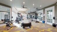 This fitness center is thoughtfully designed across a single floor, offering distinct zones that cater to a wide variety of workout styles and fitness goals. The main workout area is expansive and features bold geometric-patterned carpeting in neutral tones accented with yellow, giving the space an energetic and modern feel. Ceiling fans and recessed lighting ensure comfort throughout, while large windows along one wall flood the area with natural light and offer views to the outdoors. This zone is outfitted with TKO multi-functional strength training systems, a punching bag, medicine balls, battle ropes, a heavy-duty tire flip sled, and a selection of cardio machines including treadmills and an air bike. Full-length mirrors line portions of the walls, enhancing the sense of space and allowing exercisers to monitor their form. A dedicated functional training room adjoins the main floor, featuring built-in shelving, wall-mounted displays, and direct outdoor access through glass-paneled doors. The cycling studio is bright and airy, with vaulted ceilings, warm hardwood-style flooring, floor-to-ceiling mirrors, and generous window coverage that brings in abundant natural light. It houses several stationary cycling bikes and offers a serene, open atmosphere ideal for group or solo sessions. Glass walls and doors connect the studio to the main gym, maintaining visual continuity throughout the facility.