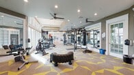 This fitness center is thoughtfully designed across a single floor, offering distinct zones that cater to a wide variety of workout styles and fitness goals. The main workout area is expansive and features bold geometric-patterned carpeting in neutral tones accented with yellow, giving the space an energetic and modern feel. Ceiling fans and recessed lighting ensure comfort throughout, while large windows along one wall flood the area with natural light and offer views to the outdoors. This zone is outfitted with TKO multi-functional strength training systems, a punching bag, medicine balls, battle ropes, a heavy-duty tire flip sled, and a selection of cardio machines including treadmills and an air bike. Full-length mirrors line portions of the walls, enhancing the sense of space and allowing exercisers to monitor their form. A dedicated functional training room adjoins the main floor, featuring built-in shelving, wall-mounted displays, and direct outdoor access through glass-paneled doors. The cycling studio is bright and airy, with vaulted ceilings, warm hardwood-style flooring, floor-to-ceiling mirrors, and generous window coverage that brings in abundant natural light. It houses several stationary cycling bikes and offers a serene, open atmosphere ideal for group or solo sessions. Glass walls and doors connect the studio to the main gym, maintaining visual continuity throughout the facility.