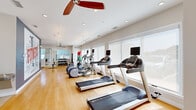 Located at 17444 Slipper Shell Way in Lewes, Delaware, this dedicated fitness center offers a bright, clean, and comprehensively outfitted workout facility on a single level. The space is finished with warm hardwood flooring throughout, contributing to an inviting yet functional atmosphere. Large windows line the walls, flooding the interior with natural light and providing views to the outdoors, while roller shades offer adjustable privacy. Ceiling fans and recessed lighting complement the airy, well-ventilated environment.

The main workout area is thoughtfully organized to accommodate a full range of exercise disciplines. Cardio equipment includes multiple treadmills, elliptical trainers, and a stationary bike, all equipped with digital displays. A rack of dumbbells and a medicine ball station support free-weight and functional training, while a multi-station cable machine with an adjacent weight bench serves strength training needs. Floor-to-ceiling mirrors in the strength zone enhance spatial awareness and allow for form monitoring. A bold fitness-themed wall mural adds motivational character to the interior. Glass doors connect to an adjacent outdoor or lounge area, and additional interior doors provide access to support rooms, making this a fully self-contained and practical fitness destination.