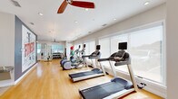 Located at 17444 Slipper Shell Way in Lewes, Delaware, this dedicated fitness center offers a bright, clean, and comprehensively outfitted workout facility on a single level. The space is finished with warm hardwood flooring throughout, contributing to an inviting yet functional atmosphere. Large windows line the walls, flooding the interior with natural light and providing views to the outdoors, while roller shades offer adjustable privacy. Ceiling fans and recessed lighting complement the airy, well-ventilated environment.

The main workout area is thoughtfully organized to accommodate a full range of exercise disciplines. Cardio equipment includes multiple treadmills, elliptical trainers, and a stationary bike, all equipped with digital displays. A rack of dumbbells and a medicine ball station support free-weight and functional training, while a multi-station cable machine with an adjacent weight bench serves strength training needs. Floor-to-ceiling mirrors in the strength zone enhance spatial awareness and allow for form monitoring. A bold fitness-themed wall mural adds motivational character to the interior. Glass doors connect to an adjacent outdoor or lounge area, and additional interior doors provide access to support rooms, making this a fully self-contained and practical fitness destination.