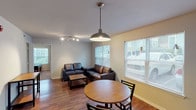 This single-floor residence offers a practical and comfortable layout designed for everyday living. The combined living and dining area serves as the heart of the home, featuring warm hardwood-style flooring, a round dining table bathed in natural light from multiple large windows with blinds, and a living space anchored by dark leather seating and a low-profile coffee table. Track lighting overhead complements the statement pendant light above the dining area. The kitchen is efficiently arranged with warm wood cabinetry, black appliances including a gas range, microwave, and refrigerator, and a peninsula counter that provides additional workspace and separation from the entryway. A dedicated front entry area leads directly into the kitchen and living spaces. Two bedrooms offer carpet flooring and windows that bring in natural light, each with a clean, neutral palette. One bedroom includes a built-in desk, adding a functional workspace. Two full bathrooms are finished with wood-look flooring, white tiled tub-shower combinations, and vanities with white countertops, providing a bright and clean aesthetic. Overall, the space presents a straightforward, functional design with consistent finishes throughout.