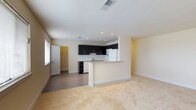 This single-floor space is designed with an open-concept layout that seamlessly connects the living area to the kitchen, creating a sense of spaciousness and easy flow throughout. The kitchen is a standout feature, outfitted with sleek dark cabinetry that contrasts sharply against the white appliances, including a full-size refrigerator. A peninsula-style breakfast bar extends from the kitchen, offering a convenient casual dining or workspace surface while maintaining the open sightlines of the room. Recessed lighting in the kitchen area provides focused, clean illumination, complementing the natural light that filters in through a window in the living space. The flooring transitions from smooth tile in the kitchen and entry zones to plush wall-to-wall carpet in the living area, adding warmth and comfort underfoot. Neutral-toned walls throughout lend a bright, airy feel that makes the space feel welcoming and versatile. A white paneled door leads to an adjacent room, suggesting additional private living quarters beyond the main open area. Overall, the space presents a clean, contemporary aesthetic well-suited for comfortable everyday living.