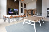 Located at 2204 Duluth Highway 120 in Duluth, Georgia, this single-story space is designed with a contemporary aesthetic that blends comfort and functionality throughout its open layout. The interior is characterized by soaring double-height ceilings in the main lounge area, where expansive floor-to-ceiling windows flood the space with natural light and offer views of the surrounding outdoor area, including a patio with umbrella seating visible beyond the glass.

The main gathering area features a curated mix of seating, including plush sectional sofas, leather lounge chairs, and a round upholstered coffee table, all anchored by layered area rugs over light hardwood-style flooring. A striking wood slat accent wall serves as a dramatic backdrop for a large multi-panel television display, enhanced by a soundbar and warm wood base cabinetry. Woven globe pendant lights in warm tones hang overhead, adding an artistic focal point.

An adjacent lounge zone transitions into a more intimate setting with a sectional sofa, dark coffee table, and rustic wooden stools, accented by black-and-white framed photography. A casual dining nook with round bar-height tables and patterned upholstered stools provides additional seating. Throughout, neutral tones, curated artwork, and thoughtful lighting contribute to a polished, welcoming ambiance.