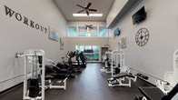 This fitness center offers a spacious, thoughtfully designed workout environment suited for a full range of exercise routines. The space features soaring ceilings with a clerestory window bank that floods the interior with natural light and provides views into an adjacent amenity area. Multiple ceiling fans maintain airflow throughout, complementing the open, airy atmosphere.

The flooring is a durable dark rubber surface ideal for high-traffic fitness use. Along one wall, a large mirror spans the width of the room, visually expanding the space and allowing users to monitor their form during workouts. A bold "WORKOUT" wall lettering and coordinating abstract artwork add motivational, contemporary flair to the neutral gray and soft teal color palette.

Cardio equipment includes treadmills, ellipticals, and stationary bikes arranged along one side of the room, while the opposite side is dedicated to a robust selection of white-framed strength training machines, including cable systems, a lat pulldown, leg press, and adjustable benches with free weights. Wall-mounted flat-screen televisions are positioned for easy viewing from both the cardio and strength training zones. A large decorative clock and curated wall art complete the polished, modern aesthetic of this well-appointed fitness facility.