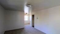 This single-floor space presents a clean, straightforward layout with a neutral color palette throughout. The main living area features wall-to-wall carpeting that adds a sense of warmth underfoot, while bright white walls create a light and open atmosphere. A ceiling fan with an integrated light fixture is positioned in an adjacent area near a window, providing both ventilation and ambient lighting. Natural light enters through the window, contributing to the overall brightness of the space. A smoke detector is mounted on the ceiling of the primary area, reflecting attention to safety. An open doorway leads to what appears to be a closet or utility area, offering practical storage or functional space. The layout is straightforward and open, allowing for flexible use and easy navigation between areas. The overall design is simple and functional, with a clean, move-in ready aesthetic. The absence of clutter and the consistent use of neutral tones throughout make the space feel welcoming and adaptable to a variety of furnishing styles and uses.