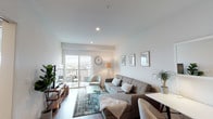 This single-floor residence offers a thoughtfully designed layout that balances functionality with contemporary style. The living room serves as the heart of the space, featuring warm wood-look flooring, recessed lighting, and crisp white walls that create a bright, airy atmosphere. A large sliding glass door floods the room with natural light and opens onto a private balcony, offering outdoor views. The space comfortably accommodates a sectional sofa, accent chair, glass coffee table, and an entertainment console, making it well-suited for both relaxing and entertaining.

The kitchen is outfitted with floor-to-ceiling light grey cabinetry, sleek hardware, white quartz countertops, and a textured tile backsplash. Stainless steel appliances—including a refrigerator, gas range, built-in microwave, and dishwasher—complete the modern aesthetic. Under-cabinet lighting adds a refined finishing touch.

The bedroom is a tranquil retreat with soft neutral tones, dual windows that invite natural light, and access to a walk-in closet. The bathroom features a clean, modern vanity with a quartz countertop, a large mirror, open shelving for storage, and a shower-tub combination enclosed by a white curtain, all set on wood-look flooring.