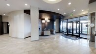 This contemporary lobby presents a polished and professional atmosphere, thoughtfully designed to make a strong first impression. The space is defined by a striking curved dark wood ceiling feature that anchors the central seating area, creating a visual focal point that draws the eye inward. Recessed lighting throughout the ceiling is complemented by a set of elegant pendant drum lights suspended over the lounge area, casting a warm, inviting glow.

The seating area is furnished with modern upholstered chairs arranged around a small accent table, set against a decorative wall panel featuring circular metallic design elements. A fireplace surround adds a refined touch to this corner of the space. The floors are finished in large-format light-toned tiles that reflect the ambient lighting, contributing to a bright and open feel.

Floor-to-ceiling glass entry doors along the perimeter allow generous natural light to pour in while offering clear sightlines to the exterior. Structural columns with clean white finishes integrate seamlessly into the design without interrupting the spacious flow. Overall, the lobby conveys a sleek, corporate aesthetic balanced with approachable warmth.