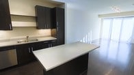 This single-floor space showcases a contemporary design aesthetic defined by clean lines and a sophisticated contrast of dark and light elements. The kitchen is a standout feature, equipped with rich, dark espresso-toned cabinetry that extends from base to upper units, offering generous storage capacity. Stainless steel hardware accents each cabinet door, adding a polished, modern touch. The countertops are finished in a crisp, light-toned surface that provides a striking contrast against the dark cabinetry. A substantial kitchen island with a matching light countertop serves as both a functional workspace and a natural divider between the kitchen and the adjoining living area. An undermount stainless steel sink is integrated into the perimeter countertop, complementing the kitchen's cohesive design. Dark hardwood-style flooring flows throughout, grounding the space with warmth and continuity. The open-plan living area beyond the kitchen is bright and airy, benefiting from what appears to be floor-to-ceiling windows that invite abundant natural light. The overall ambiance is sleek, minimalist, and well-appointed.