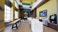 This dynamic communal space is thoughtfully designed to accommodate a variety of social and leisure activities within a single, open floor plan. The interior immediately draws attention with its bold color palette, combining deep teal, chartreuse yellow, and warm wood tones throughout. Soaring ceilings with coffered detailing and recessed lighting lend an airy, upscale feel to the main gathering area.

A long white bar counter with contemporary bar seating anchors the central space, adjacent to a wet bar station featuring a stainless steel sink, under-counter refrigeration, and a large wall-mounted television set against a distinctive geometric yellow-textured wall. This same textured wall treatment recurs throughout, adding visual depth and cohesion.

A shuffleboard table provides a focal recreational element near floor-to-ceiling windows that flood the space with natural light and offer exterior views. A dedicated lounge area features yellow patterned armchairs arranged around an oval coffee table, paired with a wood media console housing board games and a wall-mounted TV.

Private booth-style nook seating with individual tables and built-in power outlets caters to those seeking a quieter setting. A separate seating alcove with upholstered banquette, wall-lit wood paneling, and whimsical artwork creates an intimate entertainment corner. Decorative starburst wall sculptures accent the teal columns, reinforcing the space's playful, modern character.