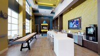 This dynamic communal space is thoughtfully designed to accommodate a variety of social and leisure activities within a single, open floor plan. The interior immediately draws attention with its bold color palette, combining deep teal, chartreuse yellow, and warm wood tones throughout. Soaring ceilings with coffered detailing and recessed lighting lend an airy, upscale feel to the main gathering area.

A long white bar counter with contemporary bar seating anchors the central space, adjacent to a wet bar station featuring a stainless steel sink, under-counter refrigeration, and a large wall-mounted television set against a distinctive geometric yellow-textured wall. This same textured wall treatment recurs throughout, adding visual depth and cohesion.

A shuffleboard table provides a focal recreational element near floor-to-ceiling windows that flood the space with natural light and offer exterior views. A dedicated lounge area features yellow patterned armchairs arranged around an oval coffee table, paired with a wood media console housing board games and a wall-mounted TV.

Private booth-style nook seating with individual tables and built-in power outlets caters to those seeking a quieter setting. A separate seating alcove with upholstered banquette, wall-lit wood paneling, and whimsical artwork creates an intimate entertainment corner. Decorative starburst wall sculptures accent the teal columns, reinforcing the space's playful, modern character.