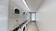 This single-floor laundry facility is designed for practicality and efficiency, providing a well-organized space for communal laundry use. The room features a continuous row of stacked commercial-grade washing machines and dryers lining one wall, offering ample capacity for multiple users. Each unit is white and modern, equipped with digital control panels for ease of use. The space is finished with light wood-effect flooring that is easy to maintain, while the walls are painted in a clean, neutral tone that reinforces the bright and tidy atmosphere. Overhead, a long LED strip light runs the length of the ceiling, ensuring the room is well-lit throughout. A window fitted with white horizontal blinds at the far end of the room allows for natural light to supplement the artificial lighting, adding to the airy feel of the space. The layout is streamlined and functional, with a clear walkway running the full length of the machines for easy access. Overall, the facility presents a hygienic, well-maintained environment designed to accommodate the laundry needs of a shared or multi-unit setting efficiently.