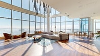 This sophisticated two-floor space offers an exceptional blend of modern architecture and breathtaking coastal scenery. The main floor is generously proportioned, featuring dramatic floor-to-ceiling curtain-wall glazing that frames panoramic waterfront, marina, and cityscape views on multiple sides. Soaring double-height ceilings amplify the sense of openness, while rich hardwood flooring and crisp white walls create a refined, contemporary aesthetic throughout. A sculptural pendant light installation cascades from above, serving as a striking focal point in the main living area, which is anchored by a large sectional sofa, a decorative area rug, and high-top bar seating positioned to maximize the views. Access to an outdoor balcony further connects the interior to its spectacular surroundings. The gourmet kitchen is outfitted with custom floor-to-ceiling cabinetry in warm natural wood tones, dark stone countertops, and premium stainless steel appliances. The upper floor, accessed via an internal staircase with glass balustrades, features chevron-patterned light wood flooring, a cozy lounge nook, and additional bar-height seating—all overlooking the main level below. Together, the two floors deliver a seamless, light-filled environment ideal for both everyday living and sophisticated entertaining.