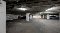 This single-level enclosed parking facility is constructed with a robust concrete structure, featuring exposed concrete ceilings, load-bearing columns, and textured concrete walls throughout. The facility accommodates a substantial number of vehicles across clearly marked parking stalls, organized into several rows separated by wide drive aisles that allow for comfortable maneuvering.

Overhead fluorescent tube lighting is distributed throughout the space, providing consistent illumination across the parking areas and drive lanes. White-painted structural columns are positioned at regular intervals, supporting the concrete ceiling above. Exposed utility runs, including pipes and conduit, are visible along the ceiling, reflecting the utilitarian nature of the facility.

Access to the facility is controlled via a gated entry and exit point equipped with a roll-up garage door, along with an additional interior gate visible in one section of the garage. Designated accessible parking stalls are marked with blue signage in multiple areas. A floor drain is visible in one of the drive lanes, supporting water management within the space. Some stalls are bordered by wooden storage enclosures, providing additional functionality for select parking spaces. The overall ambiance is functional and utilitarian, consistent with a well-established shared parking structure.