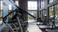 This two-story fitness center offers a well-equipped and thoughtfully designed space for a comprehensive workout experience. The facility is anchored by an impressive double-height ceiling that creates an open, airy atmosphere, complemented by expansive floor-to-ceiling windows that flood the interior with natural light and provide views of the surrounding greenery. A central staircase with modern steel and glass railings connects the two levels, serving as both a functional and architectural focal point. The lower level is outfitted with a row of cardio machines, including ellipticals and step trainers, arranged to take full advantage of the natural light streaming in from the large windows. The flooring features a light-toned surface with subtle accent detailing, adding a clean and contemporary aesthetic to the space. The upper level, accessible via the staircase, provides additional equipment and overlooks the main floor, enhancing the sense of space and scale throughout the facility. The dark-toned walls contrast effectively with the bright window frames and light flooring, giving the fitness center a sleek, modern character. Overall, the space is designed to be both functional and motivating, with an emphasis on openness, natural light, and modern design.