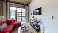 This 1-bedroom, 1-bathroom loft-style residence seamlessly merges industrial architecture with contemporary design. The living area immediately impresses with soaring ceilings that showcase exposed concrete beams, visible ductwork, and original structural elements — all hallmarks of thoughtful adaptive reuse. Light hardwood flooring runs throughout, while oversized steel-framed windows flood the space with natural light and provide access to a private balcony, ideal for outdoor relaxation.

The kitchen is outfitted with stainless steel appliances, including a range, microwave, and dishwasher, complemented by granite countertops, a mosaic tile backsplash, and a two-tone cabinet design featuring white upper cabinetry and wood-grain lower cabinets.

The bedroom continues the industrial-loft aesthetic with exposed concrete ceilings, expansive floor-to-ceiling windows with roller shades for privacy, and generous closet access. The soaring ceiling height adds an airy, open quality to the room.

The bathroom features a dark navy vanity with granite countertops, a tall linen tower cabinet for additional storage, subway tile surround, and a combination tub and shower. The overall ambiance of this residence balances raw architectural character with polished, modern finishes throughout.