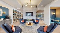 Located in Rochester, MN, this single-floor clubhouse is a generously proportioned community amenity space designed with modern aesthetics and thoughtful functionality. Light wood-look flooring flows throughout, unifying the various areas under soaring ceilings accented by striking designer light fixtures, including a sculptural brass chandelier and bold drum pendants.

The multiple living areas serve as the social heart of the clubhouse. A central lounge features a floor-to-ceiling tile-clad fireplace wall with a mounted flat-screen TV, surrounded by navy blue armchairs with orange accent pillows. Built-in open shelving in blue and natural wood tones adds character to the space. Additional lounge areas provide flexible seating arrangements, including cozy nook seating, a bar area with a teal tile backsplash, and access to a stainless steel refrigerator — ideal for entertaining.

Large floor-to-ceiling windows and glass entry doors invite abundant natural light and provide views of the surrounding community. The exercise room is well-appointed, featuring cardio equipment, weight benches, kettlebells, a wall-mounted mirror, an interactive fitness display, and patterned accent walls with a slatted wood ceiling detail. A water fountain and restroom access are conveniently located nearby. Overall, the clubhouse delivers a refined, welcoming environment for residents.