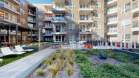 This expansive outdoor courtyard is nestled within the interior of a contemporary multi-story residential complex, characterized by a modern architectural design featuring warm taupe and rich red siding, large windows, and private balconies on every level. The courtyard is thoughtfully designed to offer a variety of recreational and relaxation zones. A generous open lawn provides flexible green space, bordered by wide stone-tile walkways that guide residents throughout the area. Alternating wood-deck tile panels add texture and warmth to the hardscape surfaces. Raised planter beds with steel-gray surrounds showcase ornamental grasses, succulents, ground cover, and seasonal plantings, creating a layered, naturalistic landscape. A dedicated activity area features a ping-pong table, a dining-height table with white chairs, vibrant orange Adirondack chairs, and a large-scale outdoor chess set, offering engaging communal spaces. Stylish lounge chairs are positioned near additional planters for relaxed sunbathing. A sculptural metallic art installation adds a distinctive focal point. The entire courtyard benefits from open-sky natural light and the enveloping presence of the surrounding building, creating a sheltered yet airy atmosphere ideal for both social gatherings and quiet outdoor enjoyment.