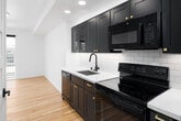 This single-floor unit showcases a cohesive modern design defined by light hardwood flooring that flows seamlessly throughout every room, creating a warm and unified aesthetic. The hallway entry leads past interior doors fitted with sleek black hardware, connecting the living spaces in an efficient and intuitive layout.

The kitchen is a standout feature, equipped with dark shaker-style cabinetry accented by brushed gold hardware, a white subway tile backsplash, a undermount sink with a matte black faucet, and a full suite of black appliances. Recessed lighting above the kitchen enhances functionality while contributing to the contemporary atmosphere.

The open living area is bright and airy, benefiting from a large sliding glass door that opens directly onto a private balcony, offering views of the surrounding urban skyline. Natural light fills the room, amplifying the clean white walls and minimalist finishes.

The bathroom continues the dark-and-light design language, featuring a dark vanity with gold drawer pulls, a white quartz countertop, an undermount sink with a matte black faucet, and a rounded rectangular mirror illuminated by a gold bar light fixture. A narrow window provides natural ventilation.

Overall, the unit presents a polished, contemporary aesthetic with thoughtful material choices throughout.