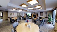 This single-floor communal space functions as both a dining and social lounge area, thoughtfully designed to accommodate group gatherings and everyday meals. The room is generously proportioned, with multiple round wooden tables distributed throughout, each surrounded by cushioned chairs upholstered in a soft floral or patterned fabric that adds a residential, comfortable feel.

A defining architectural feature of the space is the ceiling, which is lined with a series of wood-framed recessed light panels arranged in a grid pattern. These fixtures provide warm, even illumination throughout the room, contributing to an inviting ambiance.

The walls are adorned with framed botanical and nature-themed artwork, adding a calm, decorative touch. Along one wall, built-in wooden shelving units house a flat-screen television alongside books and decorative items, creating a cozy media corner. A large window at the rear allows natural light to filter in, while an indoor plant nearby enhances the welcoming atmosphere.

The neutral carpeted flooring and muted wall tones provide a cohesive, understated backdrop. Table centerpieces, including seasonal decorations and small floral arrangements, contribute personal charm to the overall setting.