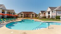 Located at 2500 McArthur Landing Cir in Fayetteville, North Carolina, this outdoor amenity space offers a well-appointed community retreat designed for relaxation and socializing. At the heart of the space is a generously sized, freeform swimming pool with crystal-clear blue water, surrounded by a broad concrete sundeck lined with teal lounge chairs and vibrant orange market umbrellas — ideal for enjoying the sunny North Carolina climate. The pool area is enclosed by a sleek black iron fence, providing both security and a polished aesthetic, with gated access points and clearly posted safety signage. Adjacent to the pool, a covered cabana-style structure offers shaded respite and additional gathering space. A dedicated outdoor lounge and social area features a raised deck platform furnished with comfortable seating, wooden picnic tables, and string lighting overhead, creating an inviting atmosphere for casual gatherings. The surrounding grounds are meticulously maintained with lush, manicured green lawns that extend between the pool and the residential buildings. The buildings themselves display a classic brick and siding exterior with pitched rooflines, balconies, and well-kept landscaping, contributing to the community's cohesive and welcoming character throughout.