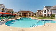 Located at 2500 McArthur Landing Cir in Fayetteville, North Carolina, this outdoor amenity space offers a well-appointed community retreat designed for relaxation and socializing. At the heart of the space is a generously sized, freeform swimming pool with crystal-clear blue water, surrounded by a broad concrete sundeck lined with teal lounge chairs and vibrant orange market umbrellas — ideal for enjoying the sunny North Carolina climate. The pool area is enclosed by a sleek black iron fence, providing both security and a polished aesthetic, with gated access points and clearly posted safety signage. Adjacent to the pool, a covered cabana-style structure offers shaded respite and additional gathering space. A dedicated outdoor lounge and social area features a raised deck platform furnished with comfortable seating, wooden picnic tables, and string lighting overhead, creating an inviting atmosphere for casual gatherings. The surrounding grounds are meticulously maintained with lush, manicured green lawns that extend between the pool and the residential buildings. The buildings themselves display a classic brick and siding exterior with pitched rooflines, balconies, and well-kept landscaping, contributing to the community's cohesive and welcoming character throughout.