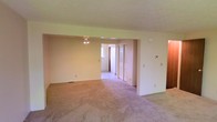 This two-floor space presents a straightforward and functional layout, characterized by its open, interconnected living areas finished with plush wall-to-wall carpeting throughout. The main living space is generously proportioned, offering an open feel enhanced by neutral cream-toned walls that create a bright, welcoming atmosphere. A ceiling fan is centrally positioned, contributing both to air circulation and ambient lighting within the space.

An open archway connects the primary living area to an adjacent room, which features a natural light source through a window, further brightening the interior. This secondary space includes two interior panel doors, suggesting access to closets or additional rooms, adding practical storage and organizational options. The consistent carpeting and color scheme flow seamlessly between areas, reinforcing a cohesive aesthetic throughout.

A thermostat is visibly mounted on the wall, indicating climate control capability. The understated, move-in ready condition of the space offers flexibility for a variety of interior design approaches. With its open layout, neutral finishes, and multi-floor configuration, this property provides a versatile foundation well-suited to a range of living arrangements.