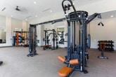 Located at 2–14 S Clayton St in Lawrenceville, GA, this fitness center is a thoughtfully designed, single-floor facility offering a comprehensive workout environment. The space is characterized by soaring ceilings and an open floor plan that creates a sense of expansiveness, allowing ample room between equipment stations for comfortable use.

Natural light floods the interior through large, floor-to-ceiling windows along the perimeter walls, providing views of the surrounding outdoor environment and contributing to an energizing atmosphere. The neutral-toned carpeted flooring, warm beige walls, and recessed lighting combine to create a clean, modern aesthetic throughout.

The strength training area is well-appointed with a variety of Matrix-brand cable machines, multi-functional cable systems, selectorized weight machines, and free-weight accessories including kettlebells and medicine balls displayed on a dedicated rack. Large wall-mounted mirrors in this section enhance the sense of space and allow users to monitor their form.

The cardio zone, positioned along the windowed wall, features a row of Matrix treadmills, Peloton-style spin bikes, and elliptical machines, all benefiting from natural light and outdoor views. Decorative textured wall panels between structural columns add a refined design touch, while mounted televisions provide entertainment options. The overall ambiance is polished, functional, and welcoming.