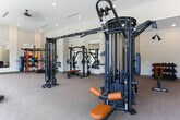 Located at 2–14 S Clayton St in Lawrenceville, GA, this fitness center is a thoughtfully designed, single-floor facility offering a comprehensive workout environment. The space is characterized by soaring ceilings and an open floor plan that creates a sense of expansiveness, allowing ample room between equipment stations for comfortable use.

Natural light floods the interior through large, floor-to-ceiling windows along the perimeter walls, providing views of the surrounding outdoor environment and contributing to an energizing atmosphere. The neutral-toned carpeted flooring, warm beige walls, and recessed lighting combine to create a clean, modern aesthetic throughout.

The strength training area is well-appointed with a variety of Matrix-brand cable machines, multi-functional cable systems, selectorized weight machines, and free-weight accessories including kettlebells and medicine balls displayed on a dedicated rack. Large wall-mounted mirrors in this section enhance the sense of space and allow users to monitor their form.

The cardio zone, positioned along the windowed wall, features a row of Matrix treadmills, Peloton-style spin bikes, and elliptical machines, all benefiting from natural light and outdoor views. Decorative textured wall panels between structural columns add a refined design touch, while mounted televisions provide entertainment options. The overall ambiance is polished, functional, and welcoming.