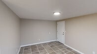 This single-floor space offers a clean, functional layout spread across several open rooms, a kitchen, and two bathrooms. Throughout the unit, neutral greige wall tones and large-format stone-look ceramic tile flooring create a cohesive and low-maintenance aesthetic.

The open living areas are bathed in natural light through multiple white-framed windows that offer views of the surrounding outdoors. Flush-mount ceiling fixtures provide consistent overhead lighting, while crisp white baseboards frame each room neatly. The rooms are generously proportioned, offering flexible use as living, dining, or sleeping spaces.

The kitchen is equipped with warm honey-toned wood cabinetry, marble-patterned countertops, a four-burner electric range with overhead ventilation hood, and a full-size refrigerator. White subway tile backsplash accents the cooking area, adding a classic finishing touch.

The first bathroom features floor-to-ceiling beige ceramic wall tile surrounding a full soaking tub and shower combination, complemented by a small privacy window. The second bathroom is finished with two-tone walls, a white vanity with mirror, a wall-mounted light fixture, and a frosted glass window for natural light. Both bathrooms continue the tile flooring found throughout the space, maintaining a unified design language.