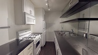 This single-floor, two-bedroom, one-bathroom residence offers a well-organized layout designed for comfortable everyday living. The galley-style kitchen is efficiently configured with white cabinetry, dark stone countertops, stainless steel double-basin sink, open metal shelving, a gas range with oven, built-in microwave, and a full-size refrigerator — all illuminated by track lighting overhead. Dark hardwood-style flooring runs consistently throughout the unit, lending a cohesive and modern aesthetic. The living area is bright and open, featuring large windows with horizontal blinds that frame views of lush outdoor greenery, allowing natural light to fill the space. A bold dark front door adds a striking contrast to the neutral interior palette. Both bedrooms are well-proportioned, each outfitted with a ceiling fan with integrated lighting. One bedroom features a distinctive two-tone accent wall with a dark lower panel, adding a contemporary design touch. The bathroom is clean and functional, with a dark vanity cabinet, a large mirror, white tile shower-tub surround, and wood-style flooring. Textured ceilings are present throughout, and the overall design balances practicality with modern finishes.