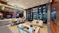 This elegantly appointed single-floor amenity space offers a seamless blend of luxury design and functional comfort. The main lounge area is anchored by a striking multi-tiered crystal chandelier that cascades from a recessed ceiling, casting a luminous glow across the room. Floor-to-ceiling built-in shelving units with dark cabinetry and teal-toned interiors line one wall, displaying curated décor and accessories that add visual depth and sophistication.

A warm wood-paneled accent wall serves as a focal point, complemented by a large flat-screen television mounted directly on it, creating an inviting media zone. Cream-toned sofas and a textured area rug define the comfortable lounge seating area, while a mirrored coffee table adds a contemporary touch.

Adjacent to the lounge, a dining area features a live-edge natural wood table surrounded by tufted upholstered chairs, set beneath a service counter and buffet station outfitted with stainless steel cabinetry and a beverage station — ideal for community gatherings or events.

Expansive floor-to-ceiling sliding glass doors connect the interior to a covered outdoor terrace with views of a resort-style pool surrounded by tropical palms. The transition between indoor and outdoor space is seamless, allowing abundant natural light to fill the interior and offering a relaxed, resort-inspired ambiance throughout.