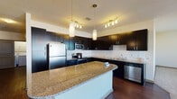 Located at 3016 Bristol Creek Dr in Morrisville, NC, this single-floor residence offers a well-designed layout that balances modern finishes with comfortable living spaces. The kitchen is a standout feature, showcasing dark espresso cabinetry that reaches toward the ceiling, granite countertops, a subway tile backsplash, and a full suite of stainless steel appliances including a refrigerator, range, microwave, and dishwasher. A large central island with pendant lighting above provides additional prep space and casual seating. Rich hardwood-style flooring flows throughout the kitchen and connecting areas. The open living area is generously sized, with plush carpeting and large windows and sliding glass doors that lead to a private balcony offering outdoor views. The bedrooms are carpeted for comfort, each featuring closets with sliding panel doors and ample natural light from well-positioned windows. The primary bathroom includes dark cabinetry with a vanity sink, a soaking tub, and access to a walk-in closet, while a secondary full bathroom mirrors the clean, modern aesthetic with a tub-shower combination and dark vanity. The overall ambiance is contemporary and functional, with a neutral color palette throughout.
