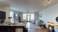 This single-floor unit offers a thoughtfully designed open-concept layout that seamlessly connects the kitchen, dining, and living areas. The living room is bright and welcoming, featuring wood-style flooring, a ceiling fan, and large windows dressed with sheer white curtains that allow generous natural light to fill the space. A wall-mounted television and a comfortable sectional sofa anchor the room, while a compact bistro-style dining nook nearby provides a casual space for meals.

The kitchen is well-equipped and functional, featuring dark espresso cabinetry, granite countertops, stainless steel appliances—including a refrigerator, range, and built-in microwave—and a double-basin sink set into a peninsula that doubles as additional prep space. Wood-style flooring continues throughout the kitchen and main living areas, adding warmth and cohesion.

The bedroom is a calm retreat with plush carpet, large windows with sheer curtains for soft natural light, and ample space for full-sized furniture. The bathroom features a granite-topped vanity with dark cabinetry, a large mirror, and wood-style flooring. A dedicated in-unit laundry closet with a full-sized washer and dryer adds everyday convenience.