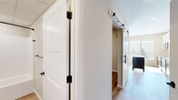 This efficiently designed one-bedroom, one-bathroom residence makes excellent use of its single-floor layout, offering a thoughtfully arranged combination of rooms finished with contemporary materials throughout. Light hardwood-style flooring flows consistently across the space, lending warmth and visual continuity from room to room.

The kitchen is a standout feature, boasting a two-tone cabinet design with crisp white upper cabinets and dark lower cabinets, complemented by a clean white countertop and stainless steel appliances including a range and microwave. A central island provides additional prep and storage space. A large window floods the kitchen with abundant natural light and offers pleasant views of the surrounding neighborhood.

The living area is bright and open, anchored by a pair of large windows that frame tree-lined rooftop views, creating an airy and inviting atmosphere. A hallway with stylish white sliding barn doors fitted with matte black hardware connects the main living areas to the private spaces, adding a modern farmhouse design element.

The bedroom is a cozy, neutral-toned retreat with clean walls and the same warm flooring found throughout. The bathroom features a dark vanity with gold-toned hardware, a white soaking tub and shower combo, and coordinating matte black fixtures, delivering a polished, spa-inspired aesthetic. The overall ambiance is clean, modern, and move-in ready.