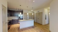 This well-appointed 1-bedroom, 1-bathroom residence offers a thoughtfully designed single-level layout that maximizes both functionality and comfort. The open-concept living and kitchen area serves as the heart of the home, anchored by a spacious center island with a granite countertop and an undermount sink, illuminated by stylish pendant lighting. The kitchen is outfitted with rich dark-espresso cabinetry, a classic subway tile backsplash, and a full suite of stainless steel appliances including a French door refrigerator with ice and water dispenser, a range, and a built-in microwave. Track lighting adds additional brightness to the cooking area, while warm hardwood-style flooring extends throughout the main living spaces, creating a cohesive and inviting atmosphere. The bedroom offers a comfortable retreat finished with plush carpeting, a ceiling fan, and a window that brings in natural light. The bathroom is well-equipped with a dark wood double vanity topped with granite, a wide mirror with overhead lighting, and a tiled soaking tub and shower combination. Multiple doors visible in the main living area suggest convenient closet and storage access, rounding out a practical and polished living space.