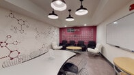 27 North is a single-floor interior space that masterfully blends scientific aesthetics with functional lounge and meeting design. The space is immediately distinguished by two striking feature walls: one finished in a light marble-like texture adorned with large-scale crimson molecular structure graphics, and another bold burgundy accent wall displaying a full periodic table of elements alongside the branded "27 North" logo — a nod to Cobalt's atomic number.

The layout accommodates multiple seating configurations. A cluster of dark upholstered armchairs surrounds a round natural wood coffee table, creating an informal gathering zone. Nearby, sleek white lounge chairs with chrome frames offer additional seating. A sculptural curved white table paired with modern wire-frame and acrylic chairs serves as a more structured meeting or work surface.

Overhead, a row of industrial-style black pendant dome lights provides focused illumination, complemented by recessed ceiling fixtures. The flooring throughout features neutral large-format tiles, grounding the space visually. Entry is through a set of bold dark double doors. A whiteboard panel along one wall adds a practical, collaborative element. The overall ambiance is dynamic, intellectually inspired, and well-suited for both casual interaction and focused discussion.
