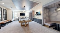 This thoughtfully designed amenity space combines entertainment, relaxation, and productivity into a cohesive, single-floor layout. The centerpiece of the space is a spacious billiards room, where a solid wood pool table anchors the room beneath a striking textured tray ceiling with an industrial-style pendant light. Multiple flat-screen TVs are mounted throughout, and tall windows allow natural light to fill the area. A built-in cabinetry unit with dark navy drawers and a clean countertop provides additional functionality, while high-top tables and bar seating line the perimeter for casual gathering.

Adjacent to the billiards area, a comfortable lounge features a large sectional sofa in soft grey, accented with teal and patterned pillows, complemented by abstract landscape artwork and warm wood flooring. A sculptural arc floor lamp in brushed gold adds an elegant touch to this relaxed seating area.

Private work alcoves offer individual desk spaces adorned with bold, artistic wallcoverings, providing a focused retreat for work or study. Glass-paneled doors connect these nooks to the broader layout, maintaining openness while preserving privacy.

A wide corridor with exposed wood ceiling beams, black iron railings, and recessed lighting ties the space together, offering views of the sunlit lounge below and leading toward additional amenity areas.