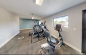 Located at 3252 SW 52nd Ave in Pembroke Park, Florida, this single-floor fitness center offers a practical and well-organized space designed to support a well-rounded workout routine. The gym area is outfitted with a variety of cardio equipment, including treadmills, ellipticals, and a stationary recumbent bike, as well as a multi-use strength training station positioned along a teal accent wall. The exercise floor features a combination of durable carpeted and concrete surfaces, providing functional zones for both machine-based and open-floor training. Natural light filters in through several windows, offering views of the surrounding outdoor area, while a wall-mounted air conditioning unit ensures a comfortable environment year-round. Flush-mounted and recessed light fixtures provide consistent overhead illumination throughout. A glass entry door allows easy access from the exterior. The adjacent bathroom is clean and modern, finished with large-format wood-look porcelain tile flooring and matching half-wall tile cladding. It includes accessible fixtures—a urinal, toilet with grab bars, and a wall-mounted sink—designed to accommodate all users. Together, the two rooms form a complete and efficiently designed fitness facility suited for community or shared-use purposes.