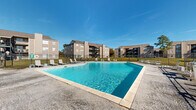 This residential complex in New Orleans offers a blend of comfort and convenience. The centerpiece is a spacious, L-shaped outdoor pool with crystal-clear turquoise water, surrounded by a concrete deck with ample lounging areas. The pool area is enclosed by a black metal fence for safety and privacy.

The property features multiple three-story apartment buildings with a neutral color scheme, balconies, and large windows that likely provide plenty of natural light to the units. The buildings have a modern architectural design with clean lines and a mix of materials including brick and siding.

Landscaping around the complex includes well-maintained lawns, mature trees, and neatly trimmed shrubs, adding to the overall aesthetic appeal. The grounds are equipped with paved walkways and outdoor seating areas, encouraging community interaction and outdoor relaxation.

Additional amenities visible include a single-story clubhouse or common area building near the pool, suggesting potential indoor recreational spaces. The property is secured with perimeter fencing, and there appears to be ample parking available for residents.

Overall, this complex offers a resort-like atmosphere with its inviting pool area and well-kept grounds, making it an attractive option for those seeking a comfortable living environment with outdoor recreational opportunities in New Orleans.
