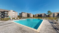 Located at 5131 Bundy Rd in New Orleans, Louisiana, this outdoor community pool area serves as a central amenity within a well-established residential complex. The generously sized swimming pool is the focal point of the space, featuring clear blue water and a classic rectangular shape with gently curved edges. Depth markers are visible along the pool's edge, and metal entry rails provide safe and convenient access.

Surrounding the pool is a broad concrete sun deck outfitted with multiple lounge chairs, offering ample space for sunbathing and relaxation. The pool area is fully enclosed by a black iron security fence with a gated entry, ensuring a secure and private environment for residents. A low-profile utility and storage structure is positioned adjacent to the pool, housing mechanical equipment and providing practical support for the amenity.

The backdrop features several three-story residential buildings with neutral earth-tone exteriors, balconies, and staircase access, framing the pool area on multiple sides. Mature trees and neatly trimmed landscaping provide greenery along the perimeter. Under the open Louisiana sky, the space benefits from abundant natural light, making it an inviting outdoor retreat. The overall setting reflects a functional and community-focused outdoor amenity designed for everyday enjoyment.