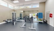 The fitness center at 25001 SW 130th Ave in Princeton, FL is a well-appointed, single-level gym designed to support a comprehensive workout experience. The space features soaring ceilings with recessed panel lighting, lending an open and airy feel throughout. Warm taupe walls are complemented by durable dark rubber flooring, creating a clean, professional aesthetic.

The main workout area is lined with large clerestory windows on multiple sides, flooding the space with natural light and offering views of the surrounding outdoor greenery and pool area. A full-length wall mirror further enhances the sense of depth and openness. Equipment is thoughtfully arranged throughout, including treadmills positioned directly in front of expansive glass doors that overlook the resort-style pool — an inviting backdrop for cardio sessions.

The strength training section is well-equipped with a multi-station cable machine, a full rack of dumbbells, adjustable benches, and a medicine ball storage rack. A dedicated cardio zone houses elliptical trainers and a recumbent cycle, with a wall-mounted TV for entertainment. The entry area features high ceilings, a cubby storage unit, and direct glass door access to the outdoor amenity area. Hand sanitizer stations and safety signage are conveniently placed throughout the facility.