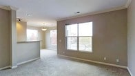 This single-floor residence offers a welcoming and functional layout designed to maximize comfort and natural light. The main living area is spacious and open, featuring plush wall-to-wall carpeting that adds warmth underfoot. Large windows dominate the walls, flooding the room with abundant daylight and providing views of the surrounding neighborhood, including mature trees and adjacent buildings. Crown molding trims the ceiling, lending a clean and polished finish to the space. A tasteful pendant light fixture is centrally positioned, providing warm overhead illumination that complements the natural light. The neutral, earthy wall tones create a calm and versatile backdrop, making the space adaptable to a variety of interior styles. A partial wall with a raised countertop separates the living area from the kitchen, creating a natural division while maintaining an open, connected feel. The kitchen area benefits from its own window, ensuring consistent brightness throughout. Ceiling vents are thoughtfully placed for efficient climate control. Overall, the space presents a clean, move-in-ready environment with classic architectural details, generous natural light, and a practical, open-concept flow that suits everyday living.