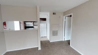 This single-floor residence offers a straightforward and functional layout designed for everyday comfort and ease of living. The open-concept design connects the living area to the kitchen, separated by a partial wall that adds subtle definition while maintaining an open feel. The living space features wall-to-wall carpet in a warm neutral tone, contributing to a cozy and understated ambiance throughout.

The kitchen is efficiently arranged with standard appliances, including a freestanding range and a full-sized refrigerator, complemented by dark wood cabinetry above and a light-colored countertop. Tile flooring in the kitchen provides a practical contrast to the carpeted living area, clearly delineating the two spaces.

A built-in wall HVAC unit is centrally positioned, ensuring climate control is both accessible and unobtrusive. The textured ceiling adds a traditional character consistent throughout the space. A doorway visible from the main living area leads to an additional room, offering private separation within the unit.

Overall, the space presents a clean, neutral palette with beige and white tones dominating the interior, making it a versatile canvas well-suited for a range of furnishing styles. The practical design and included appliances make this a move-in ready space.